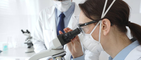 Two scientists in lab coats and safety gear conducting research, one observing through a microscope...