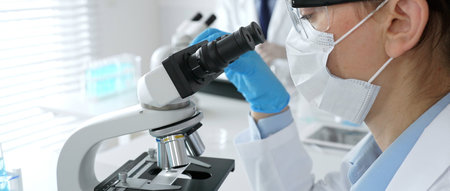 A female scientist in a blue mask and gloves is using a microscope and conducting scientific...