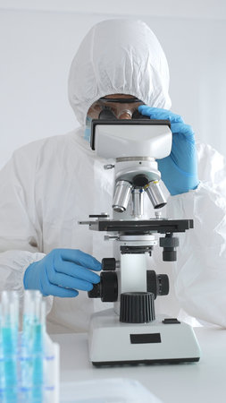 A medical virologist in protective gear examines viral samples under a microscope while conducting...