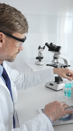 Male adult scientific researcher wearing protective eyewear, holding blue liquid test tube, working...
