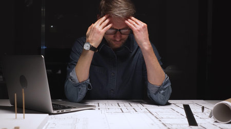 Architect or engineer working late, focusing on technical drawing with a compass, blueprint, laptop...