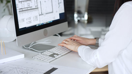 Architect working at desktop computer, checking blueprints and design plans for a new engineering...
