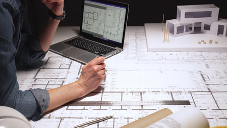 Architect planning building construction on blueprints, a modern house model nearby, using laptop...