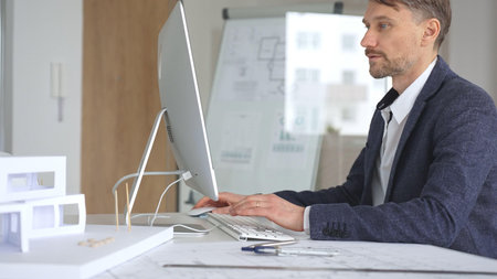Architect working on computer in office, designing new building plan with blueprint and...