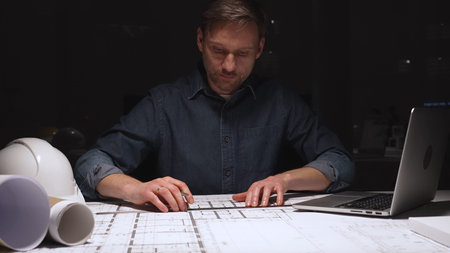 Architect working late into the night, focusing on detailed blueprints and construction plans using...