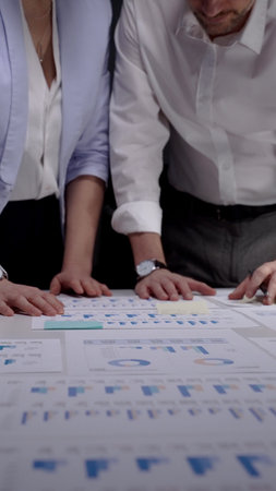 Business colleagues collaborating late at night, hands pointing at financial documents with graphs...