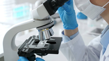 Scientific professional wearing blue protective gear examining microscope lens, performing detailed...