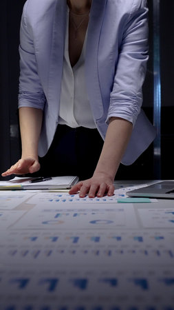 Businesswoman analyzing corporate financial data and sales growth charts on documents spread across an office desk, working late night in a professional settingの写真素材