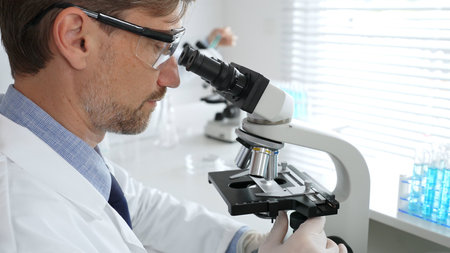 Medical scientist wearing gloves and protective glasses examining samples under a microscope in a...