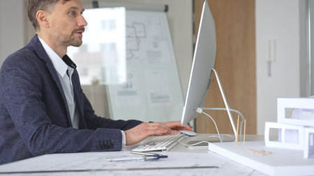 Architect working on computer in office, designing new building plan with blueprint and...