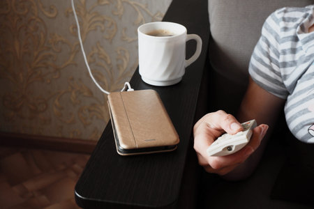 a woman switches TV channels with a remote control. Nearby, a cell phone is charged via a wire,