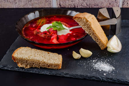 a plate of borscht is on a dark table.there are onions, garlic and some bread nearby ..の写真素材