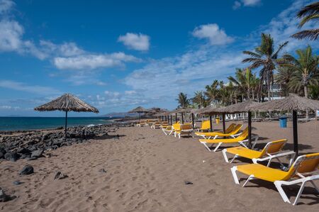 Empty beach in Canarian island Lanzarote. White sand, black lava rocks, umbrellas and yellow sun chairs. Summer holiday and tropical vacation concept.の写真素材