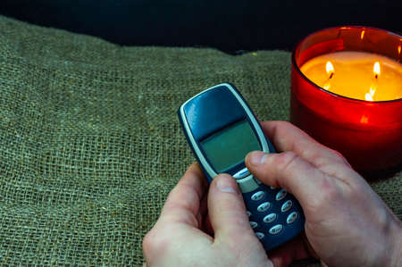 an old push-button mobile phone in elderly hands on the background of a burning candleの写真素材