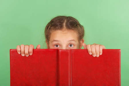 Beautiful school girl peeping from behind her red book isolated on greenの写真素材