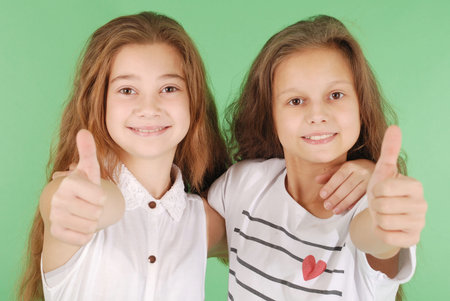 Two smiling young school girls showing thumbs up isolated on green background