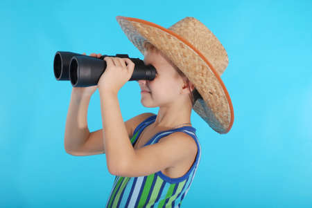 Curious boy in cowboy hat looking through binoculars. Photo on a blue backgroundの写真素材