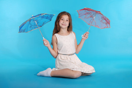 Cheerful young girl in white dress with an umbrella sitting against the blue backgroundの写真素材