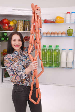 Young girl holds sausages on the refrigerator background. Beautiful young girl near the fridge.の写真素材