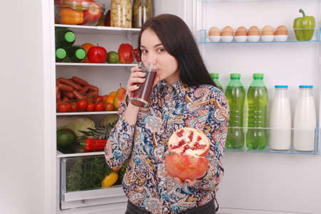 Beautiful smiling young girl holds a glass of pomegranate juice and garnet on the fridge backgroundの写真素材