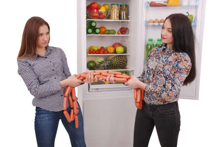 Two young girls hold sausages on the refrigerator background. Two beautiful young girls near the fridge.の写真素材