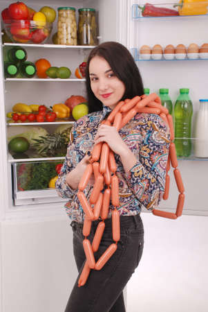 Young girl holds sausages on the refrigerator background. Beautiful young girl near the fridge.の写真素材