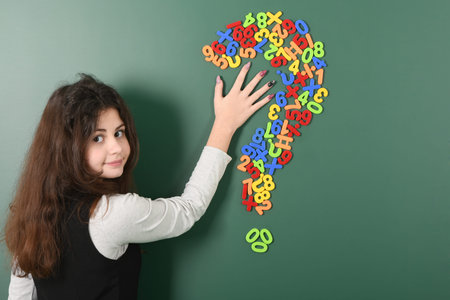 Smiling little schoolgirl on green school board. On the part of her head there is a sign of the question of magnetic lettersの写真素材