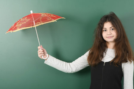 Smiling little schoolgirl on green background . Young playful girl holds a red umbrella in her handの写真素材