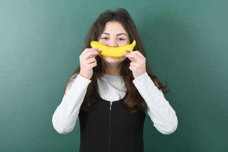 Smiling little schoolgirl on green background . Young playful girl holds a banana in his handの写真素材
