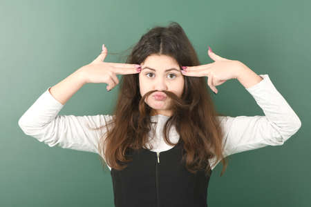 Smiling little schoolgirl on green background . Young playful girl making mock moustache using her own hairの写真素材