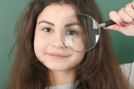 Smiling little schoolgirl on green background . Young playful girl holds a magnifying glass in his handの写真素材