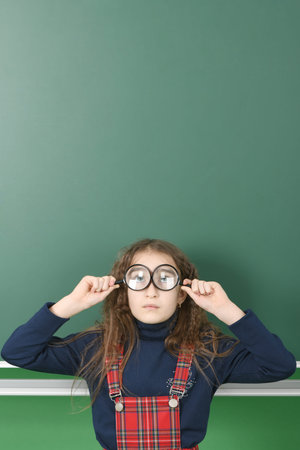 Schoolgirl near green school board. Young playful girl holds a magnifying glass in his hand. High...