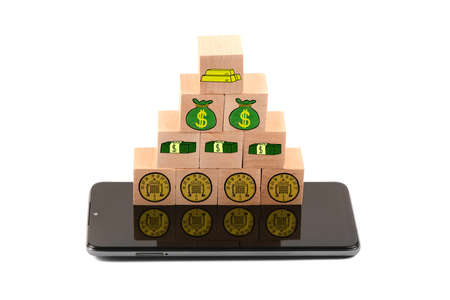 Black smartphone under wooden blocks with painted money, coin, gold, pack of bills, bags. Isolated on white background. High resolution photo. Full depth of field.の写真素材