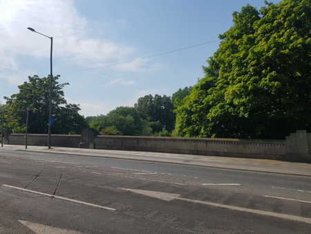 Sunlit view of the main road out of town with a stone wall lining the footpath and flourishing foliage under a bright blue summer sky.の写真素材