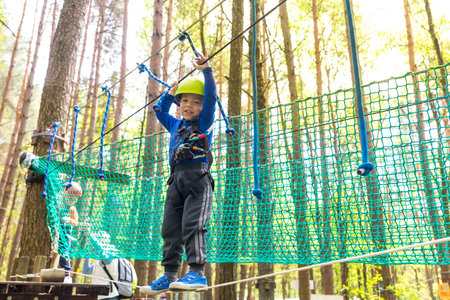 Young boy in helmet walks by rope at sunny summer day in adventure playground