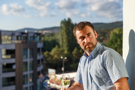 handsome guy standing on balcony in luxury hotel in Bulgaria at sunset and drinking white wine.の写真素材