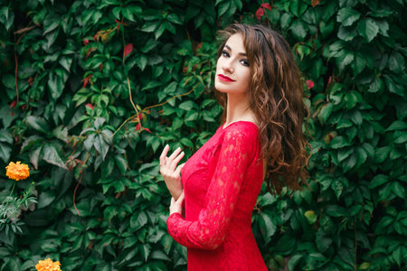 beautiful young woman in long red dress on the background of a wall with ivyの写真素材