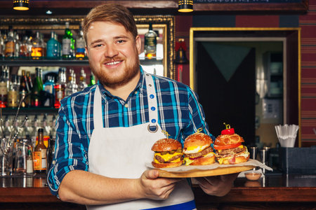 Cheerful young Chef in apron presenting a tray with different delicious burgersの写真素材