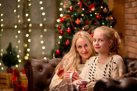christmas and family concept - mother and daughter talking and drinking tea sitting on sofa at home with Christmas treeの写真素材
