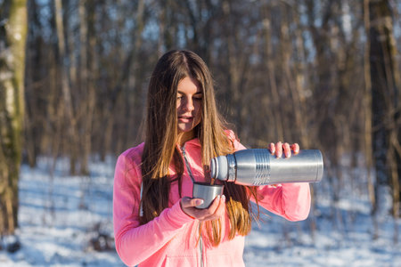 Beautiful young woman pours tea from a thermos after jogging in the winter forest.