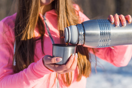 Beautiful young woman pours tea from a thermos after jogging in the winter forest. Selective focus.の写真素材