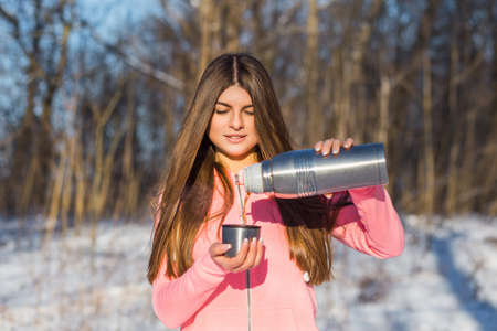 Beautiful young woman pours tea from a thermos after jogging in the winter forest. Selective focus.の写真素材