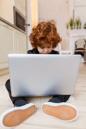 Little redhead boy uses a laptop while sitting on the floor at homeの写真素材