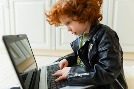 Little redhead boy uses a laptop while sitting on the floor at homeの写真素材