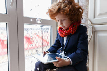 Little redhead boy uses a tablet PC sitting in his room at homeの写真素材