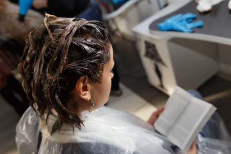 Beautiful young woman reading a book while waiting with hair dye in her headの写真素材