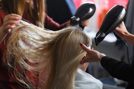 Two masters at the same time dried with hair dryers hair blonde model in beauty salon. Rear view.