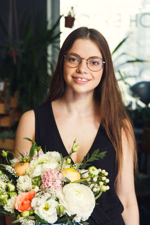 Cheerful young lady in black clothes and round glasses with bouquet of different flowersの写真素材