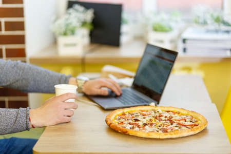 Business or study concept. Pizza stands on a table in a cafe. A person combines eating and working on a computer.の写真素材