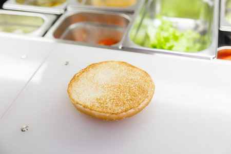 Half of the hamburger bun is on the cook's work surface. Next to it are stainless steel containers...